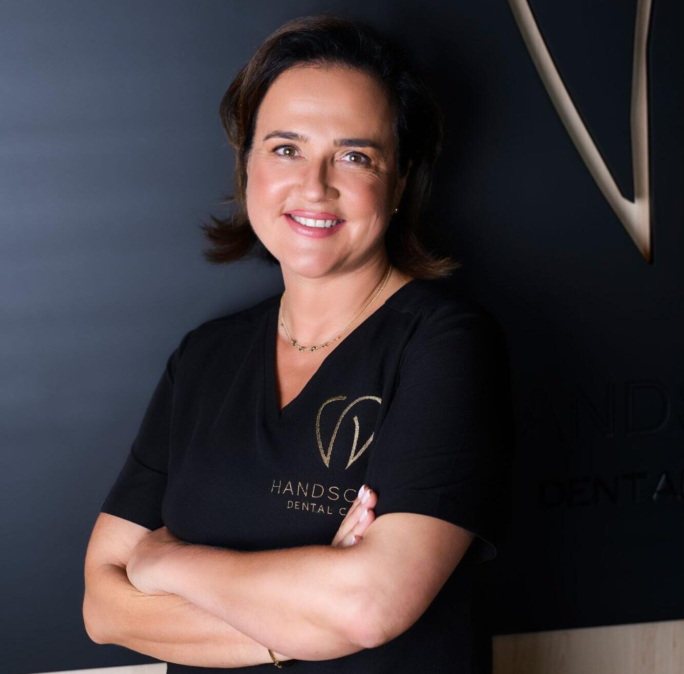 Dr Dorota Handschuh, stomatolog i właścicielka Handschuh Dental Clinic, uśmiechnięta, w czarnym uniformie medycznym, stoi z założonymi rękami na tle logotypu kliniki.