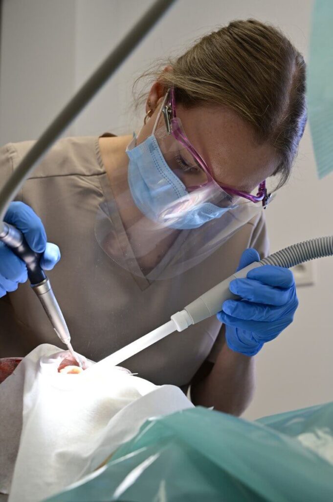 Higienistka stomatologiczna w przyłbicy, masce i rękawiczkach wykonuje zabieg oczyszczania zębów pacjenta przy użyciu specjalistycznych narzędzi.