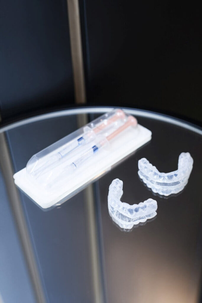 Przezroczyste nakładki termokurczliwe na zęby oraz dwie strzykawki z preparatem wybielającym na szklanym stoliku.