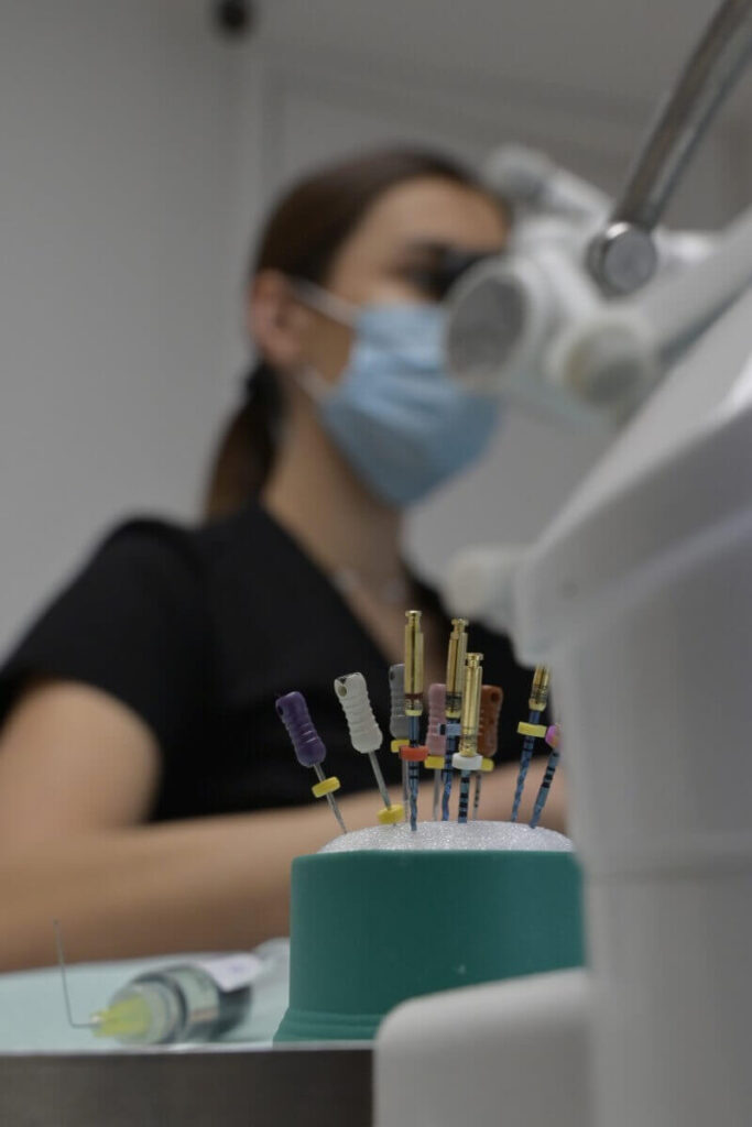 Zestaw kolorowych, precyzyjnych pilniczków endodontycznych wbitych w zieloną gąbkę; w tle rozmyty profil lekarza stomatologa w masce.