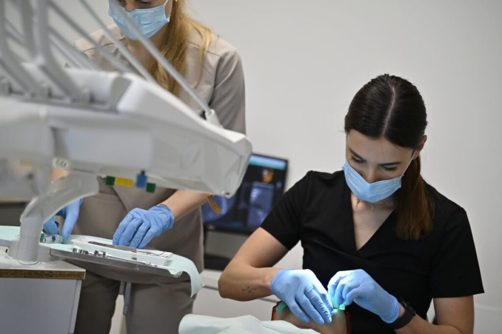 Lekarz stomatolog w czarnym uniformie i asystentka w beżowym uniformie pracują w duecie przy pacjencie; widoczne precyzyjne przygotowanie narzędzia.