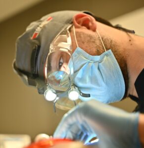 Zbliżenie na lekarza stomatologa wykonującego precyzyjny zabieg medyczny. Lekarz nosi okulary ochronne, maskę chirurgiczną oraz specjalistyczne lupy zabiegowe z oświetleniem LED, które pozwalają na pracę w dużym powiększeniu.