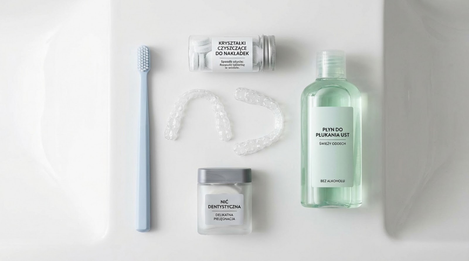 Widok z góry na akcesoria do higieny nakładek prostujących: jasnoniebieska szczoteczka, przezroczyste nakładki, kryształki czyszczące w słoiczku, nić dentystyczna oraz bezalkoholowy płyn do płukania ust na białym tle.